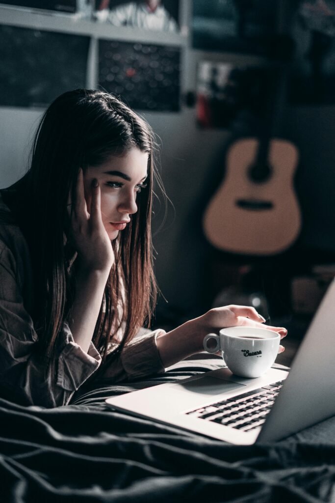 Woman lying on bed, focused on laptop with a coffee cup in hand, embracing remote work lifestyle.